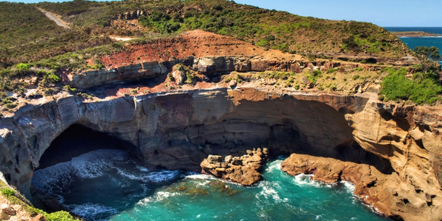  A rocky shoreline hollowed out by crashing turquoise water to form one large and two smaller caves, with scrubby red earth on the shore above. 