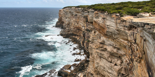  A steep cliffside with horizontally eroded layers of rock, topped with thick grass, stretching into a dark blue sea.
