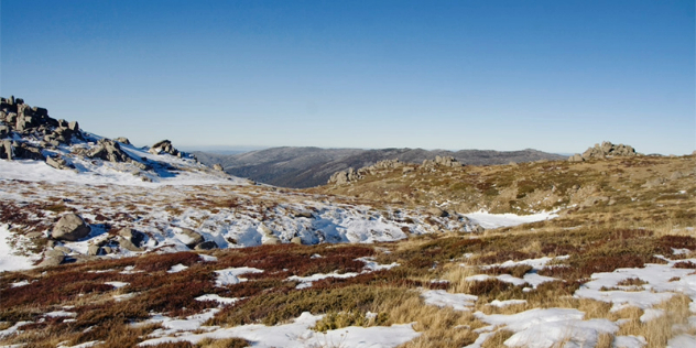  Patches of snow dotting green and yellow grassy and rocky hills that lead to a valley and mountain range in the distance.