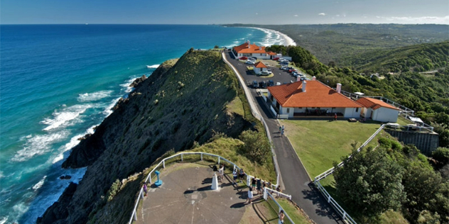  View from a lighthouse, looking down at a concrete base with tourists, a road and white sand and forested coastline far below.